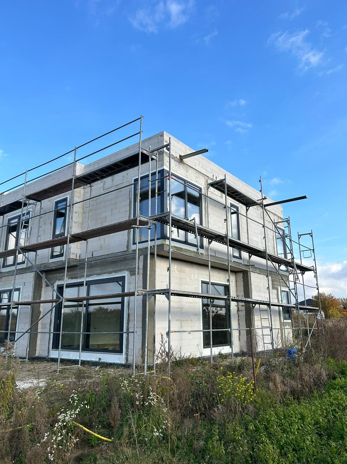 Surowy budynek z szarego betonu komórkowego w trakcie montażu okien z czarnymi ramami, otoczony rusztowaniami na tle błękitnego nieba.