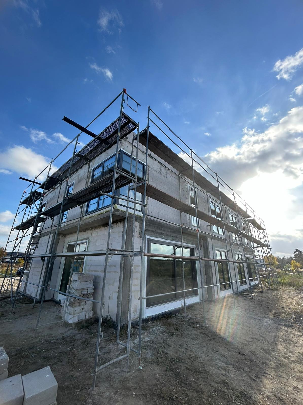 Dom w trakcie budowy z zamontowanymi oknami, otoczony rusztowaniem na tle błękitnego nieba z chmurami. Widoczne bloczki betonowe i elementy konstrukcyjne.