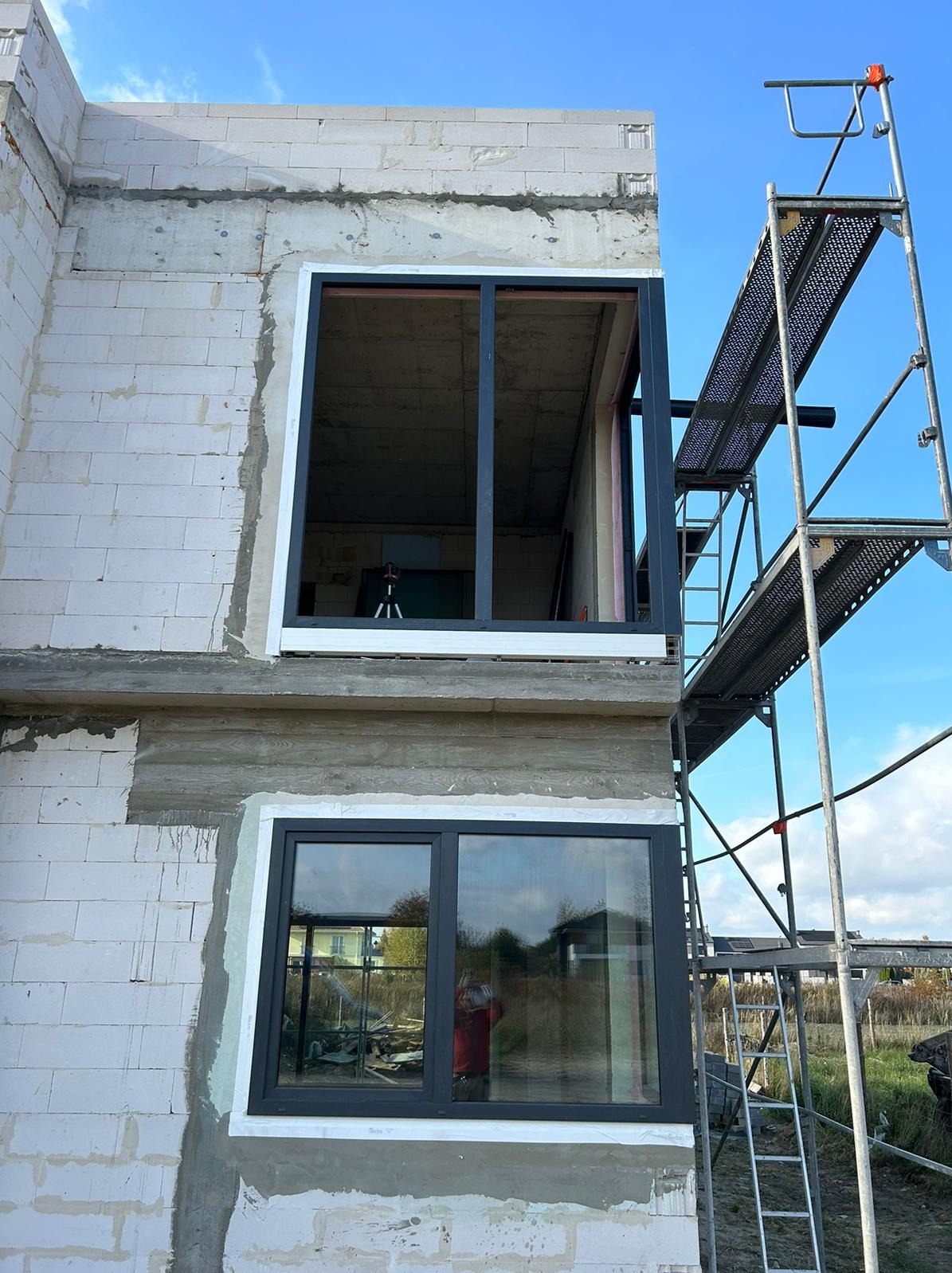 Montaż okien z czarną ramą w surowym budynku z białego bloczka. Widoczne rusztowanie i odbicie otoczenia w szybie. Dwa okna na różnych kondygnacjach.