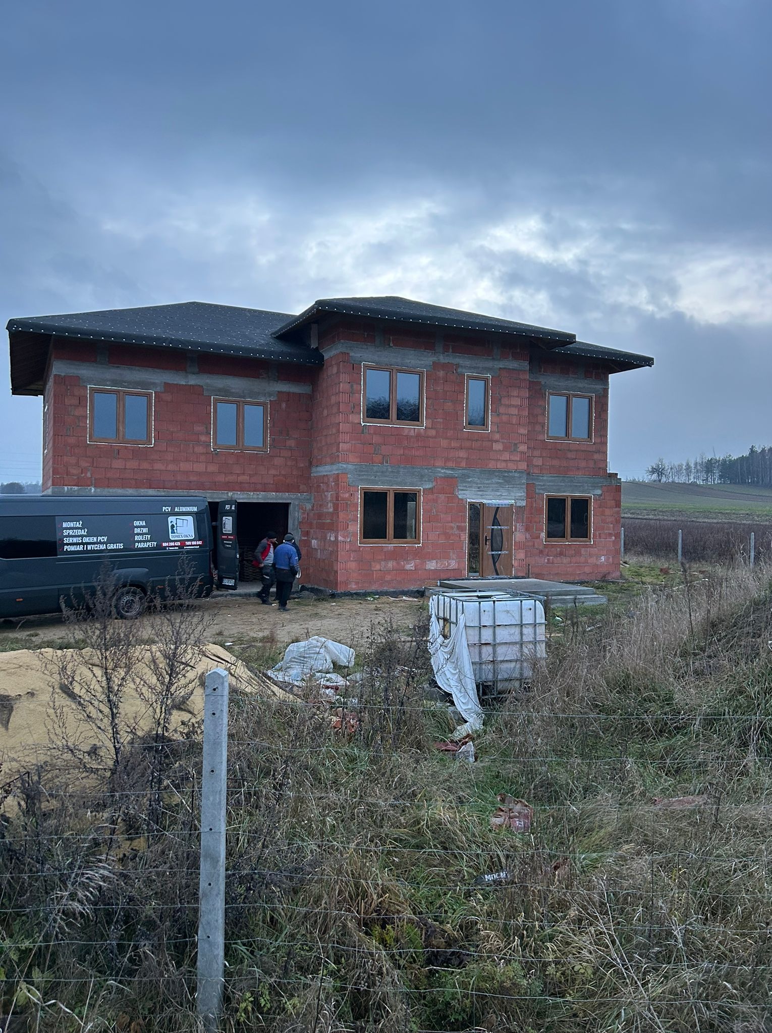 Budowa domu w stanie surowym otwartym z zamontowanymi oknami. Na pierwszym planie bus firmy montującej okna. Pochmurne niebo.