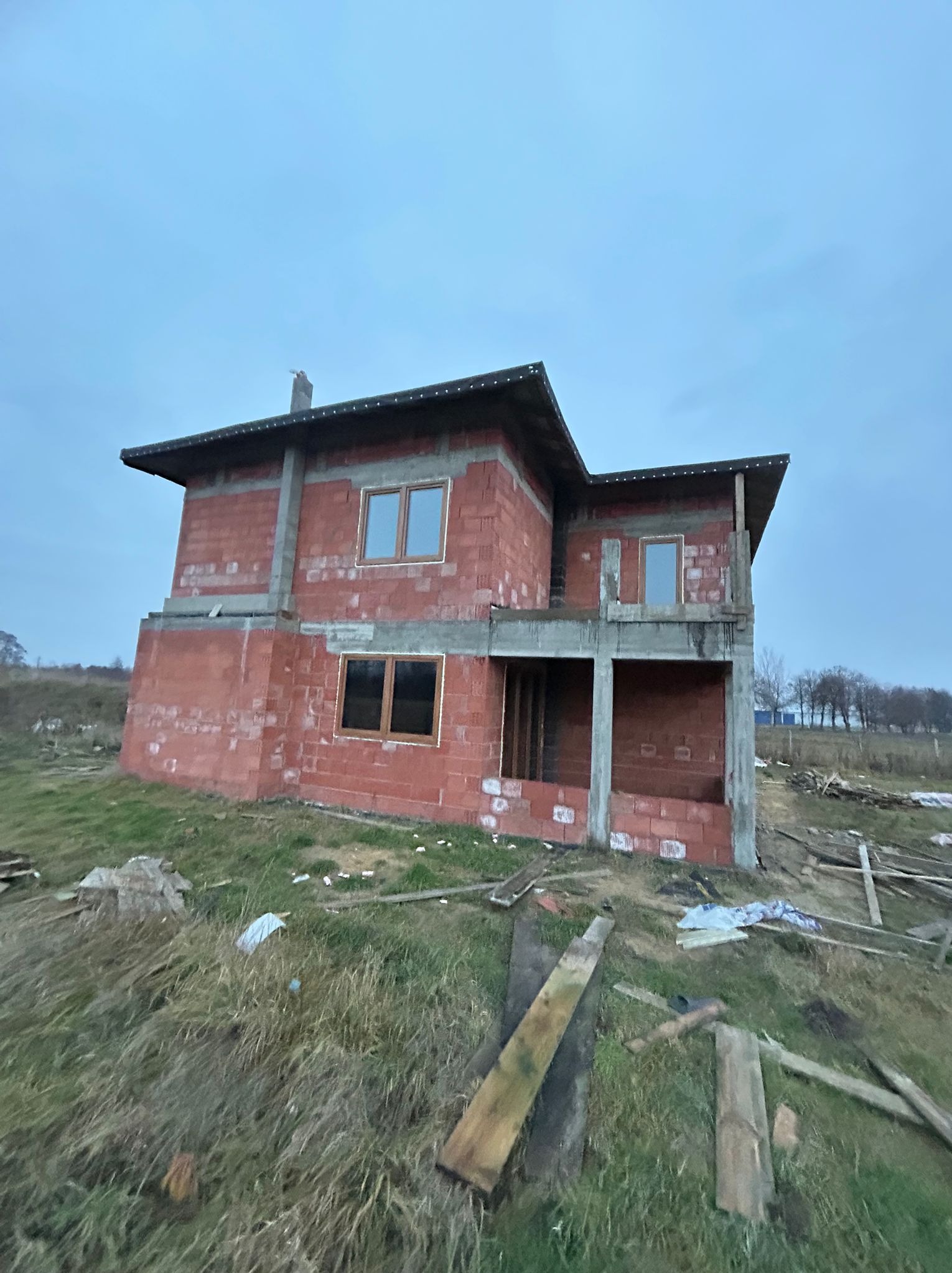 Surowy stan dwukondygnacyjnego domu z czerwonej cegły z zamontowanymi oknami, otoczonego trawą i drewnianymi deskami. Widok z dołu na tle pochmurnego nieba.