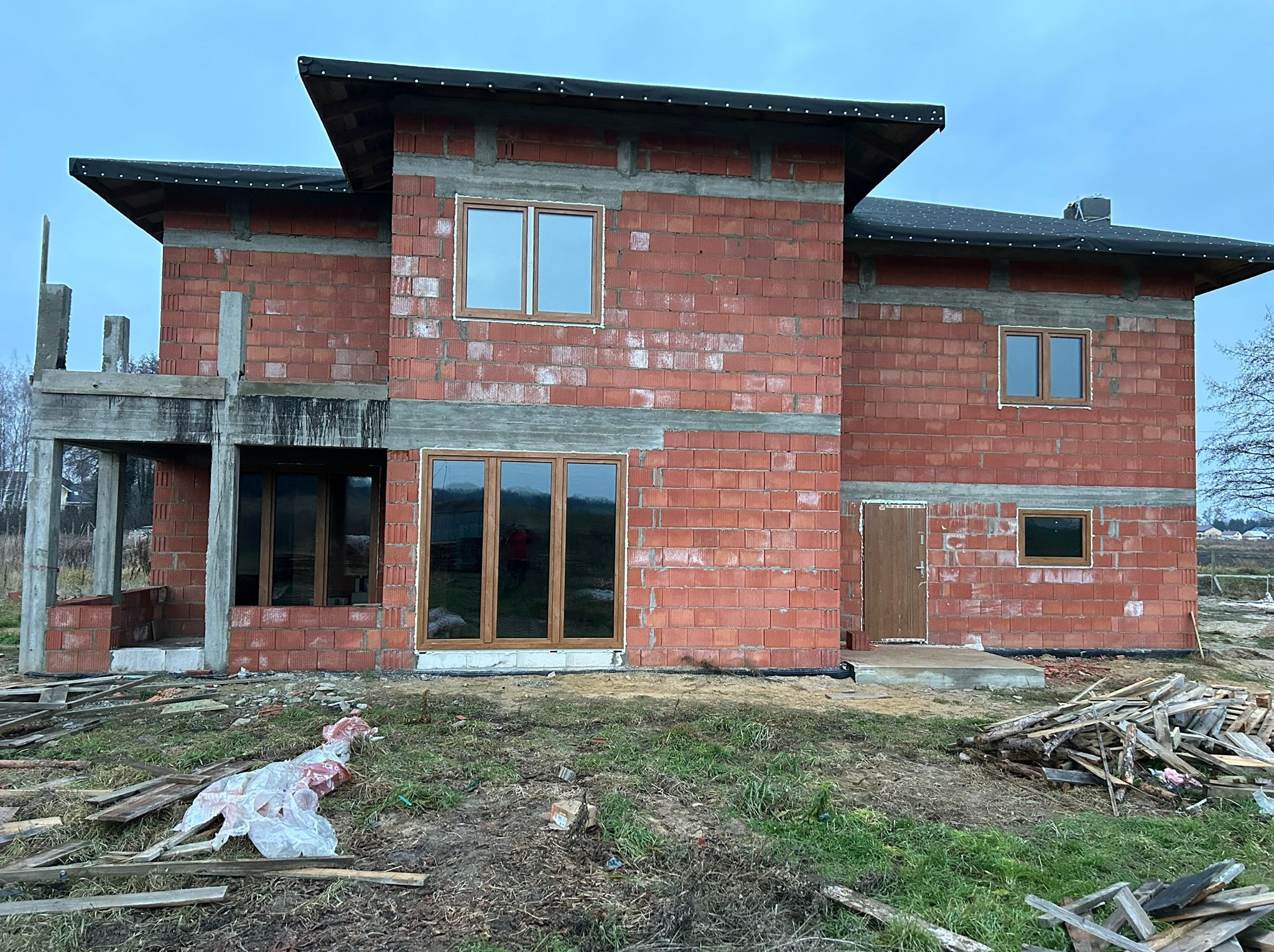Surowy stan budynku z czerwonej cegły z zamontowanymi oknami o drewnianych ramach. Widoczne niedokończone elementy konstrukcyjne i materiały budowlane na zewnątrz.