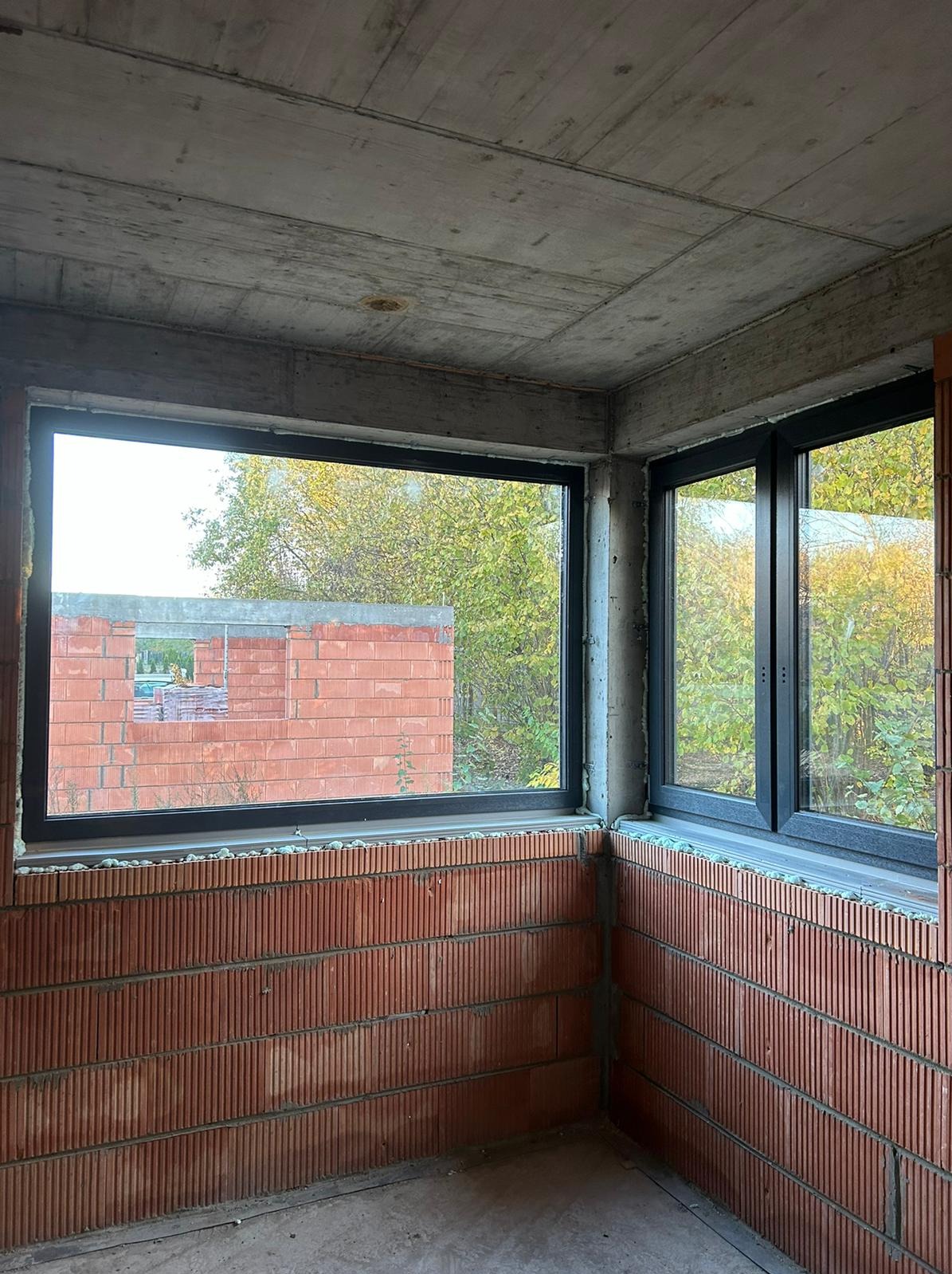 Surowy stan: czarne ramy okienne w murze z cegły dziurawki, widok na zieleń i budynek w budowie. Betonowy sufit, narożne okno.