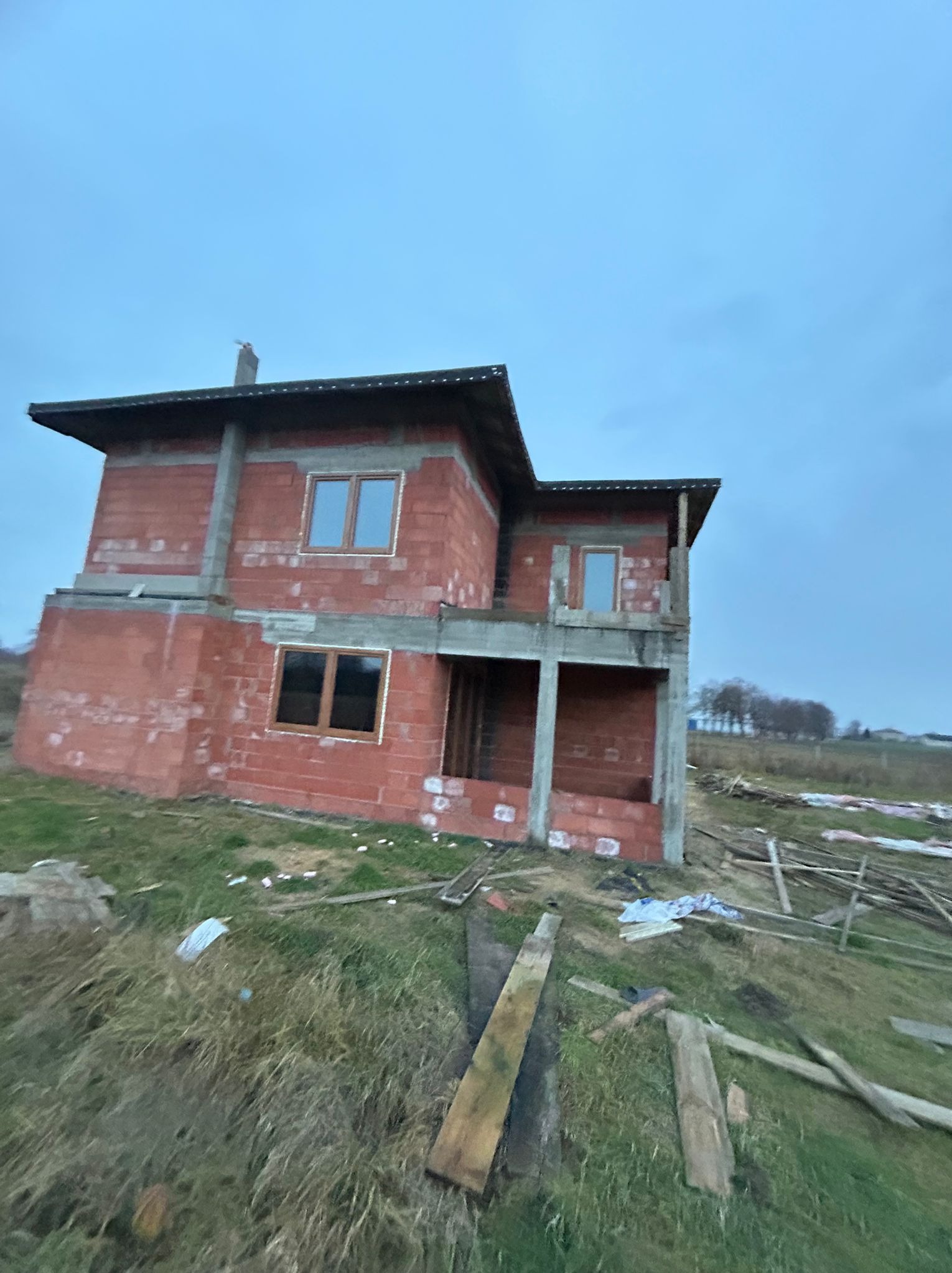 Surowy stan dwukondygnacyjnego domu z czerwonej cegły z zamontowanymi oknami z brązowymi ramami, na tle pochmurnego nieba i zarośniętej trawy.