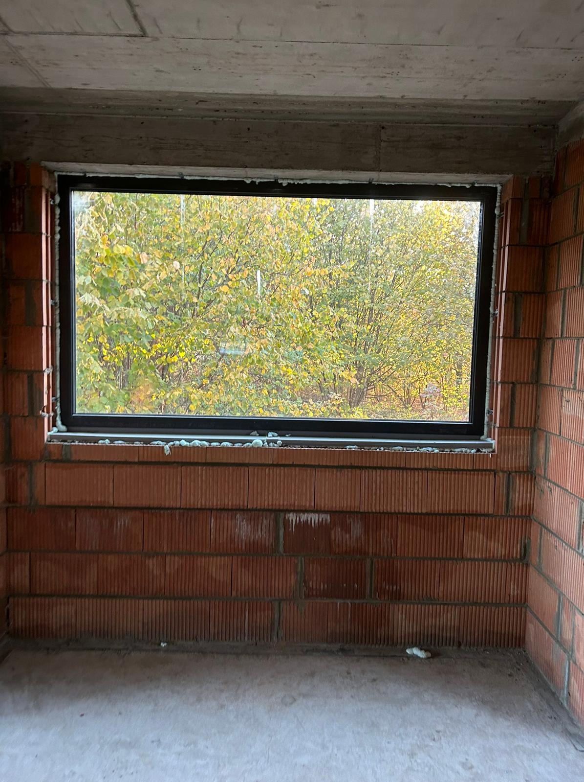 Ciemna rama okienna w surowym murze z widokiem na jesienne drzewa. Montaż okna w trakcie budowy, widoczne pianki montażowe.