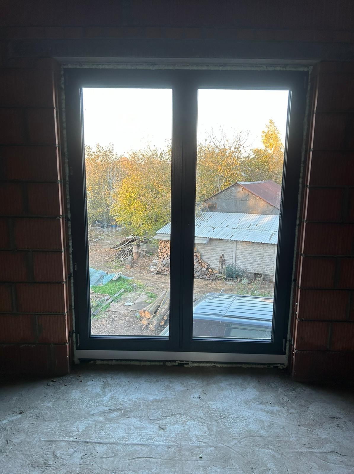 Zamontowane, dwuskrzydłowe okno w surowym murze. Widok na podwórze z drzewami i budynkiem gospodarczym. Pianka montażowa wokół ramy okiennej.
