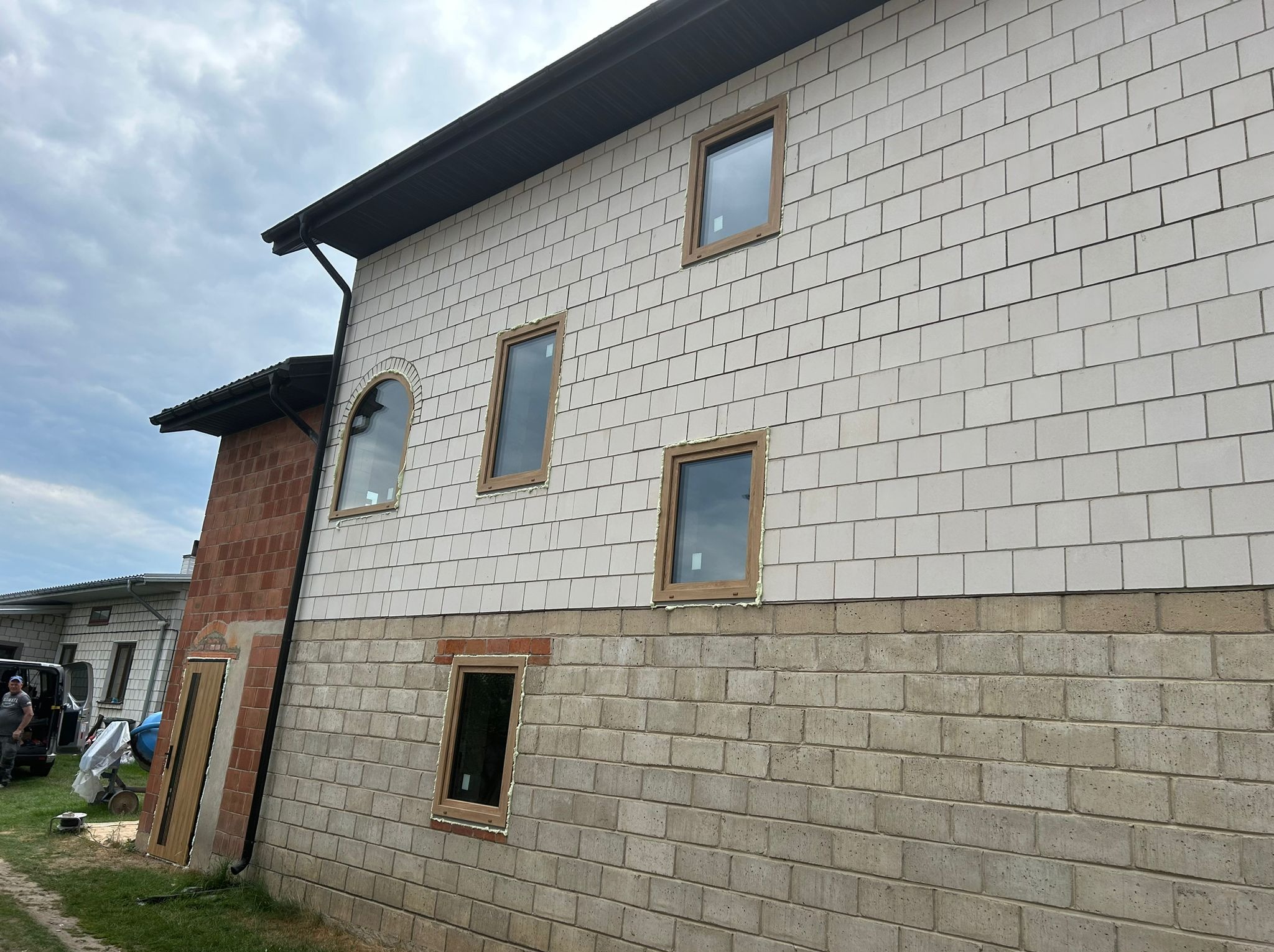 Montaż okien w nowym domu z elewacją z białej cegły i ceglanym dołem. Widoczne ramy okienne z pianką montażową. Częściowo widoczna osoba i betoniarka.