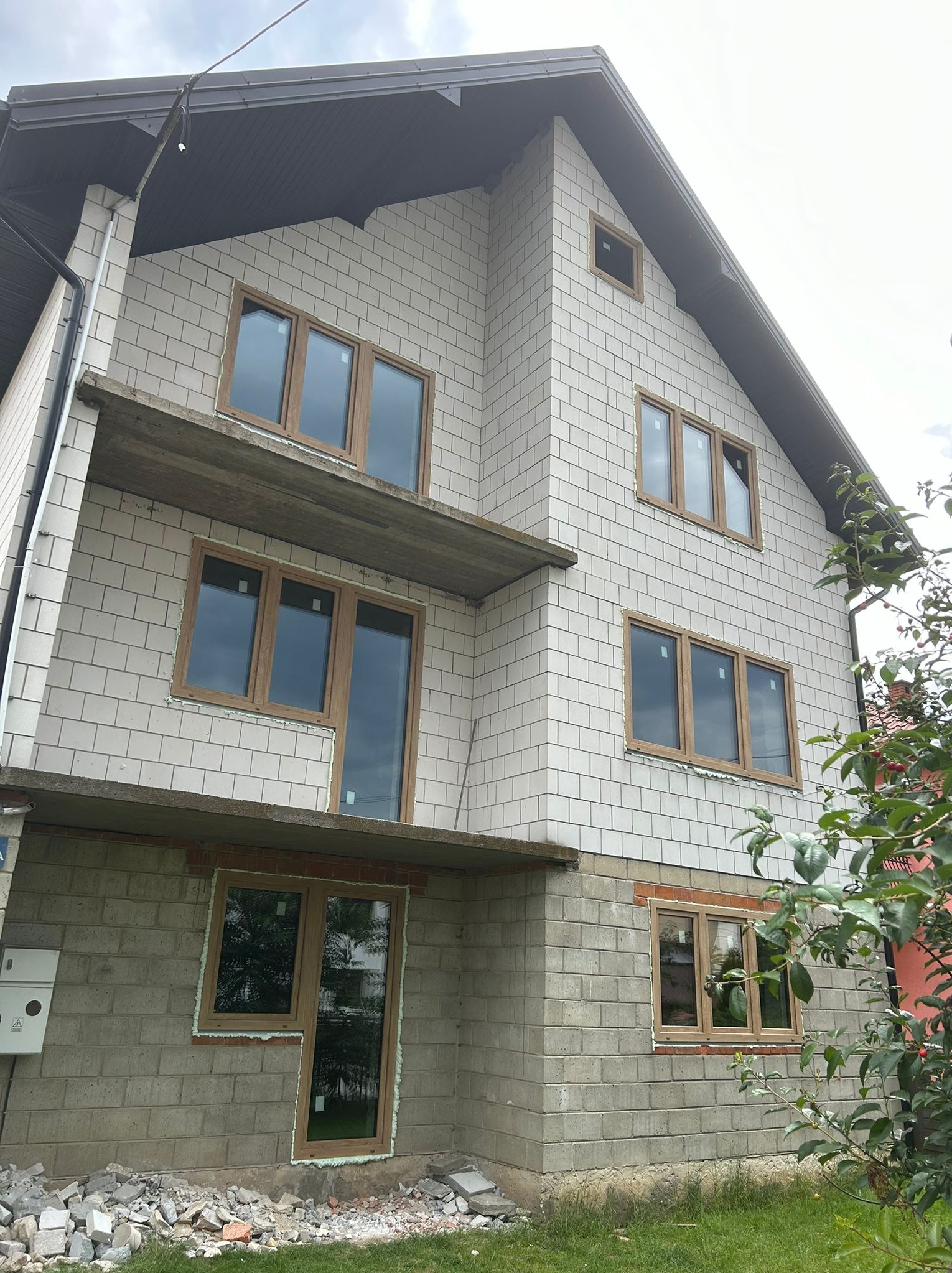 Montaż okien z brązowymi ramami w surowym budynku z białej cegły. Widoczne niedokończone elewacje i gruz. Ujęcie z dołu, pochmurne niebo w tle.