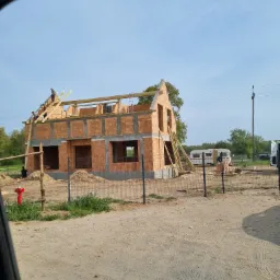 Budowa domu murowanego w trakcie realizacji: widoczne ściany z cegły, drewniana konstrukcja dachu, pracownicy na rusztowaniu, teren budowy z piaskiem i ogrodzeniem z siatki, ujęcie zza szyby...