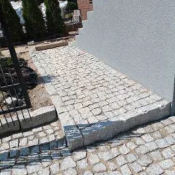 Ułożona z kostki brukowej ścieżka prowadząca do ściany budynku, widoczne obrzeża i fragment ogrodzenia.