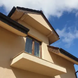 Fragment elewacji domu murowanego w Drzonowie, z widocznym oknem, balkonem i brązowym dachem na tle błękitnego nieba.