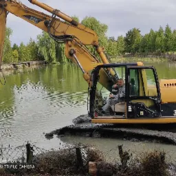 Żółta koparka CAT 315C L zanurzona w stawie podczas prac ziemnych, operator pokazuje kciuk w górę.