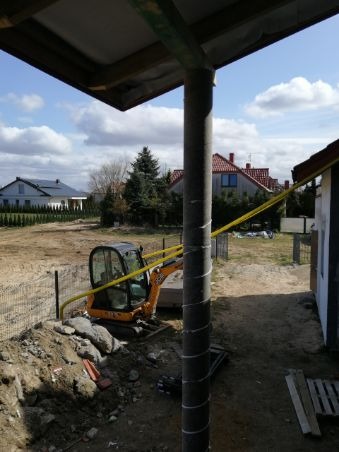 Żółta mini koparka na placu budowy, widok z zadaszonego miejsca, w tle domy jednorodzinne i częściowo ogrodzony teren.