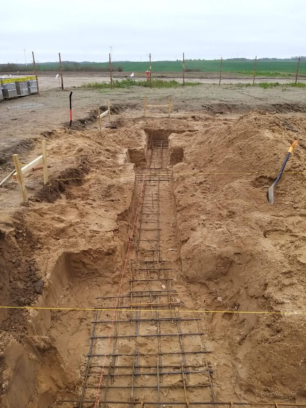 Wykop pod fundament z ułożonym zbrojeniem stalowym w ziemi, widoczna szpadel wbity w ziemię i paliki wyznaczające granice, w tle pole i turbiny wiatrowe.