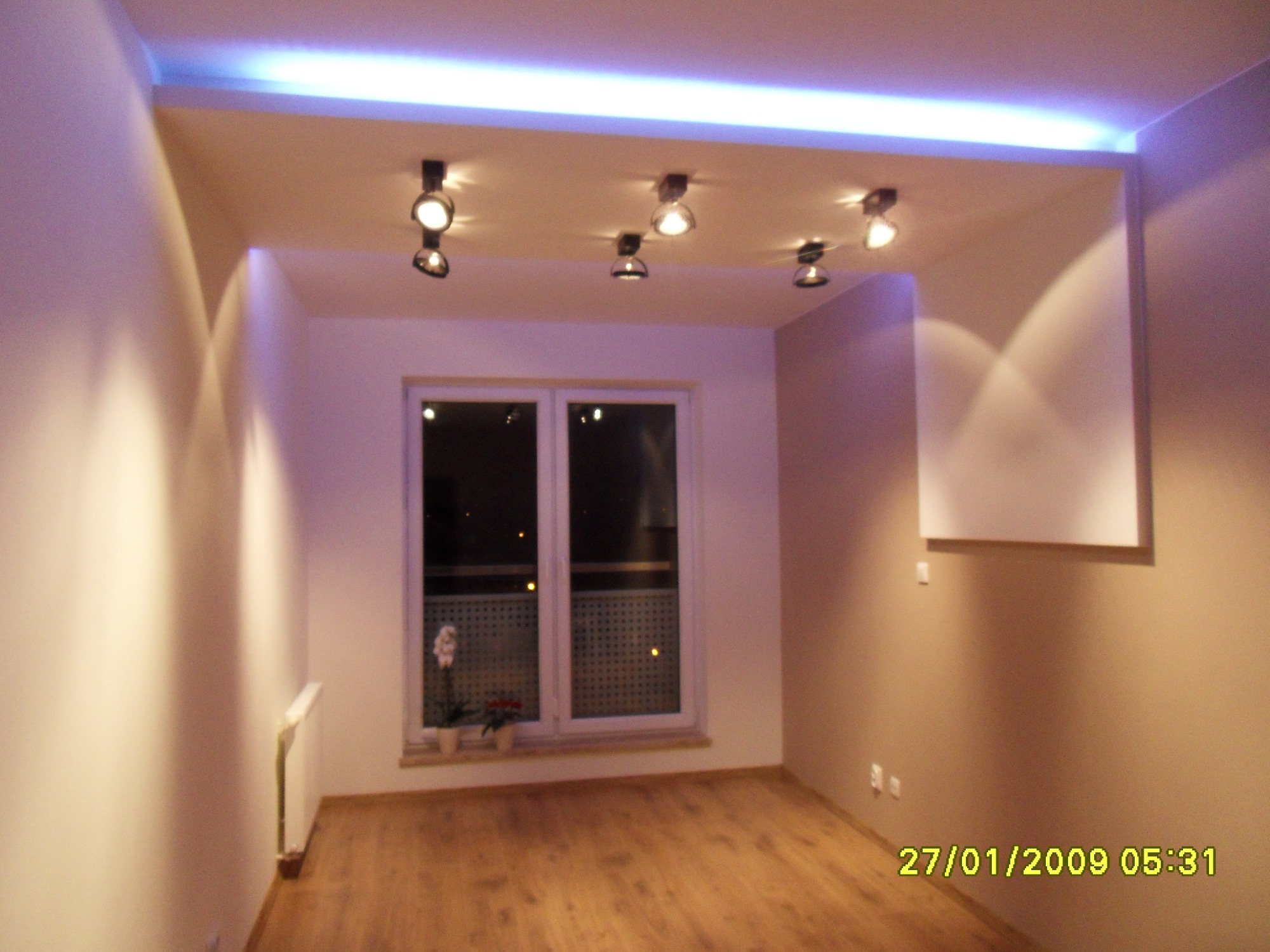 Pokój z drewnianą podłogą, białymi ścianami, oświetleniem sufitowym w postaci halogenów i listwy LED oraz oknem z widocznymi kwiatami na parapecie.