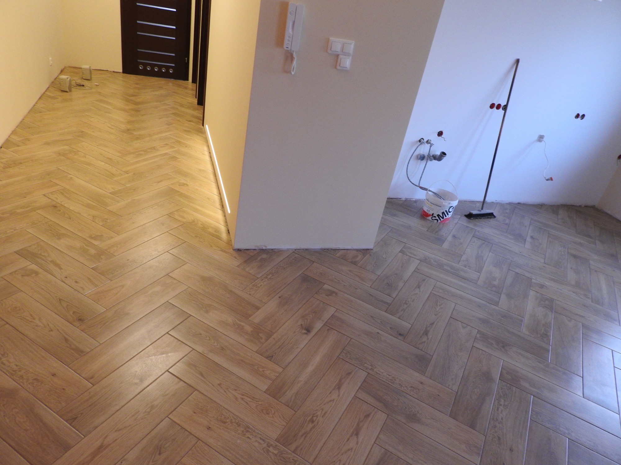 Parkiet ułożony w jodełkę na podłodze w korytarzu, widoczny fragment ściany z włącznikami i domofonem oraz przygotowane przyłącza hydrauliczne w sąsiednim pomieszczeniu.