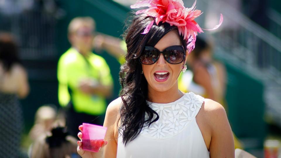 Kobieta w białej sukience z kwiatami i różowym kapeluszu, trzymająca różowy napój, uśmiechnięta podczas imprezy plenerowej.