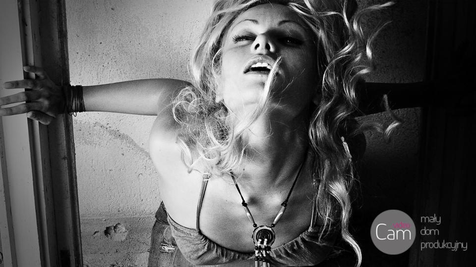 Czarno-białe, artystyczne ujęcie kobiety z rozpuszczonymi blond włosami, skierowanej twarzą ku górze, z ręką dotykającą ściany, w tle widoczne logo firmy produkcyjnej.