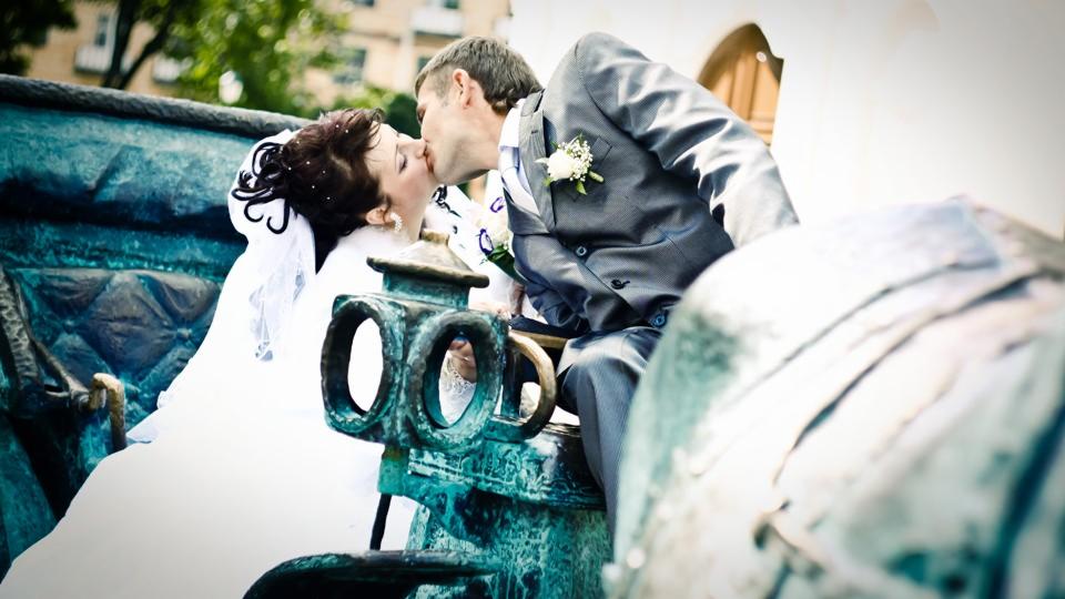 Pocałunek nowożeńców w ozdobnej, błękitnej karocy, panna młoda w białej sukni z welonem, pan młody w szarym garniturze z butonierką.