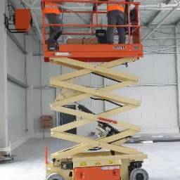 Dwóch elektryków w pomarańczowych kamizelkach na podnośniku nożycowym JLG, montujących kable pod sufitem w hali magazynowej z widoczną instalacją elektryczną.