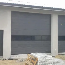 Dwie szare bramy garażowe segmentowe z oknami, zamontowane w szarym budynku, obok drzwi wejściowe, paleta z workami zaprawy budowlanej przed budynkiem.