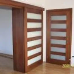 Dwa skrzydła drzwi wewnętrznych z drewnianą ramą i poziomymi, matowymi przeszkleniami. Jedno skrzydło zamontowane w ościeżnicy, drugie oparte o ścianę. Widoczny fragment podłogi i ściany.