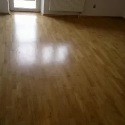 Błyszcząca, świeżo wycyklinowana podłoga parkietowa w pustym pokoju, z widocznym radiatorem i oknem w tle.