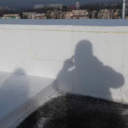 Ocieplenie dachu styropianem z widocznym cieniem osoby fotografującej, w tle zabudowa miejska.
