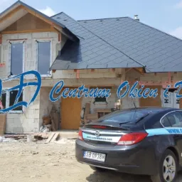 Dom w trakcie budowy z zamontowanymi oknami, widoczne logo firmy VEKA, samochód firmy Centrum Okien i Drzwi z reklamą na karoserii, w tle krajobraz górski.