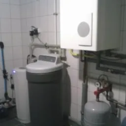 Zestaw urządzeń w kotłowni: zmiękczacz wody, zbiornik hydroforowy, piec i orurowanie na tle białych płytek ceramicznych.