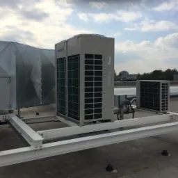 Zewnętrzne jednostki klimatyzacyjne Samsung zamontowane na metalowej konstrukcji na dachu budynku, widok na tle pochmurnego nieba i zabudowań w oddali.