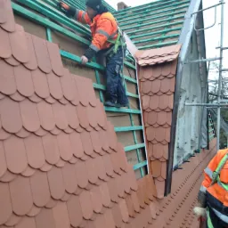 Pracownik w odblaskowej odzieży montuje dachówkę karpiówkę na nowej konstrukcji dachu z zielonych łat, widoczne rusztowanie i fragment elewacji budynku.