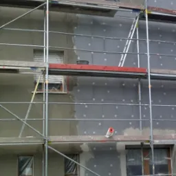 Dwukondygnacyjny dom w trakcie ocieplania elewacji. Część ściany pokryta izolacją, druga w szarym tynku. Rusztowanie, butelka wody i rolka folii na pierwszym planie.