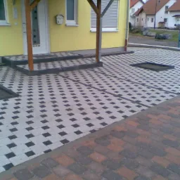 Żółty dom jednorodzinny z zadaszonym wejściem i ułożoną kostką brukową w dwóch wzorach: szaro-czarnym oraz ceglasto-szarym.