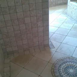 Wykończona łazienka z beżowymi płytkami kwadratowymi na ścianach, podłogą z dużych płytek oraz dekoracyjną mozaiką w kształcie koła w rogu pomieszczenia.