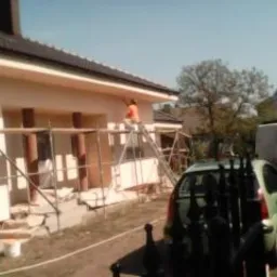 Prace elewacyjne przy domu jednorodzinnym: malowanie elewacji z rusztowania, widoczne kolumny i fragment dachu, samochód dostawczy w tle. Słoneczny dzień.