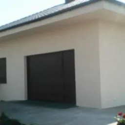 Jasny, otynkowany budynek garażowy z brązową bramą segmentową i małym oknem. Przy oknie stoi drabina. Czerwone kwiaty w dole kadru. Ujęcie z boku, lekko z dołu.