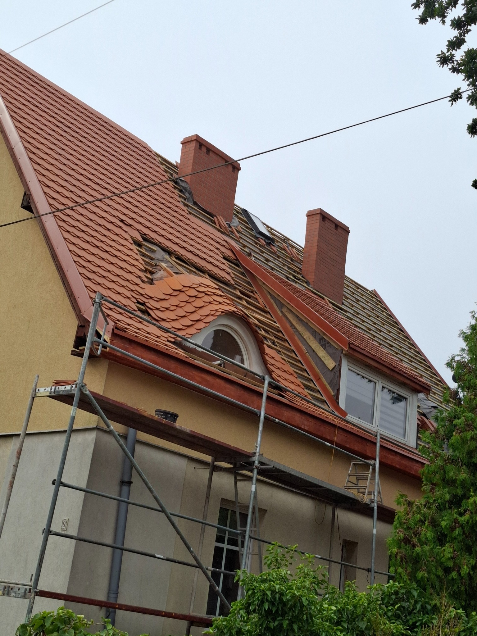 Dom jednorodzinny z częściowo zdjętym czerwonym dachem ceramicznym, widoczne rusztowanie i komin, etap prac dekarskich.
