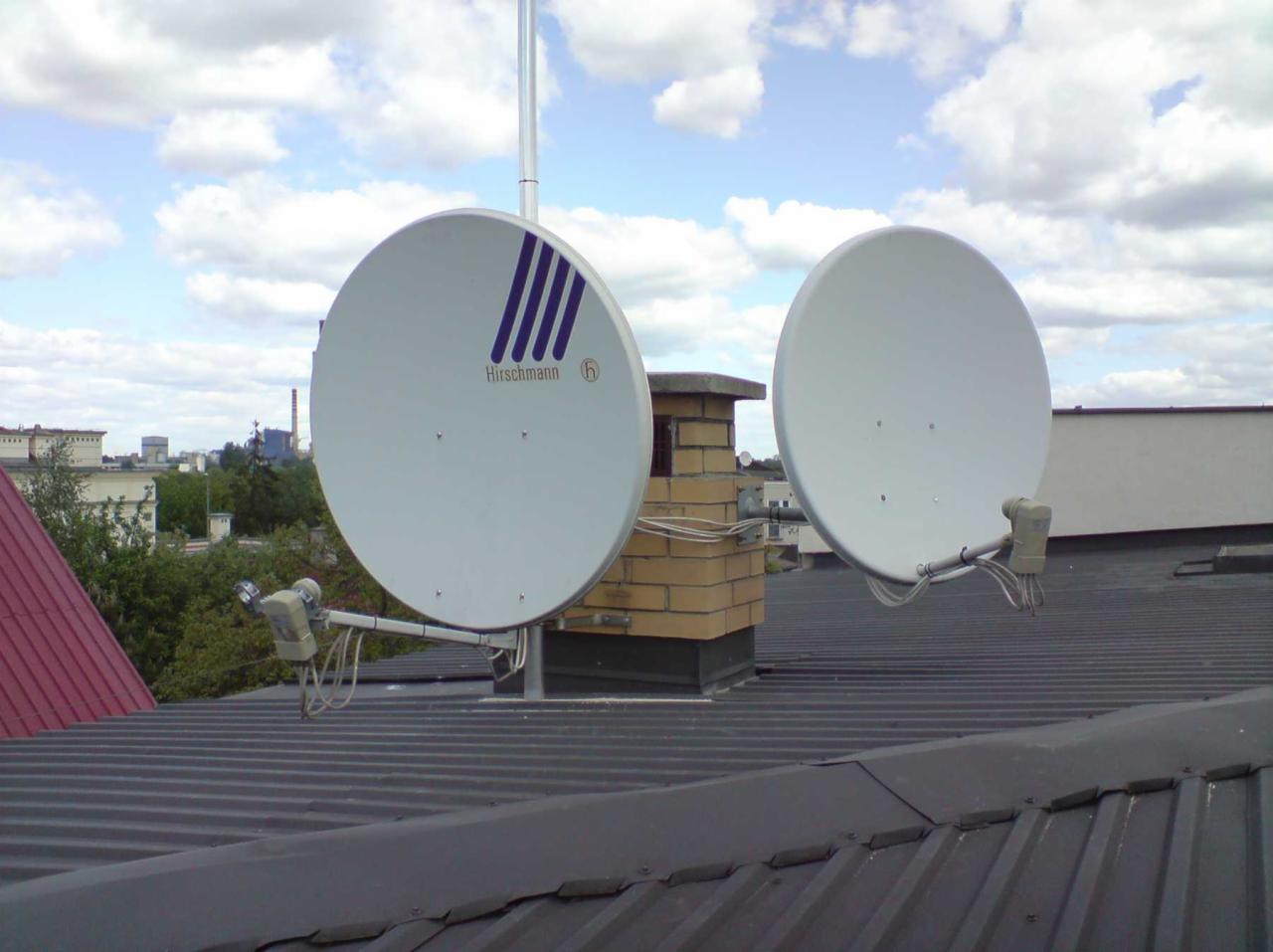 Dwie białe anteny satelitarne marki Hirschmann zamontowane na szarym, blaszanym dachu, z ceglanym kominem pośrodku i widokiem na miasto w tle.