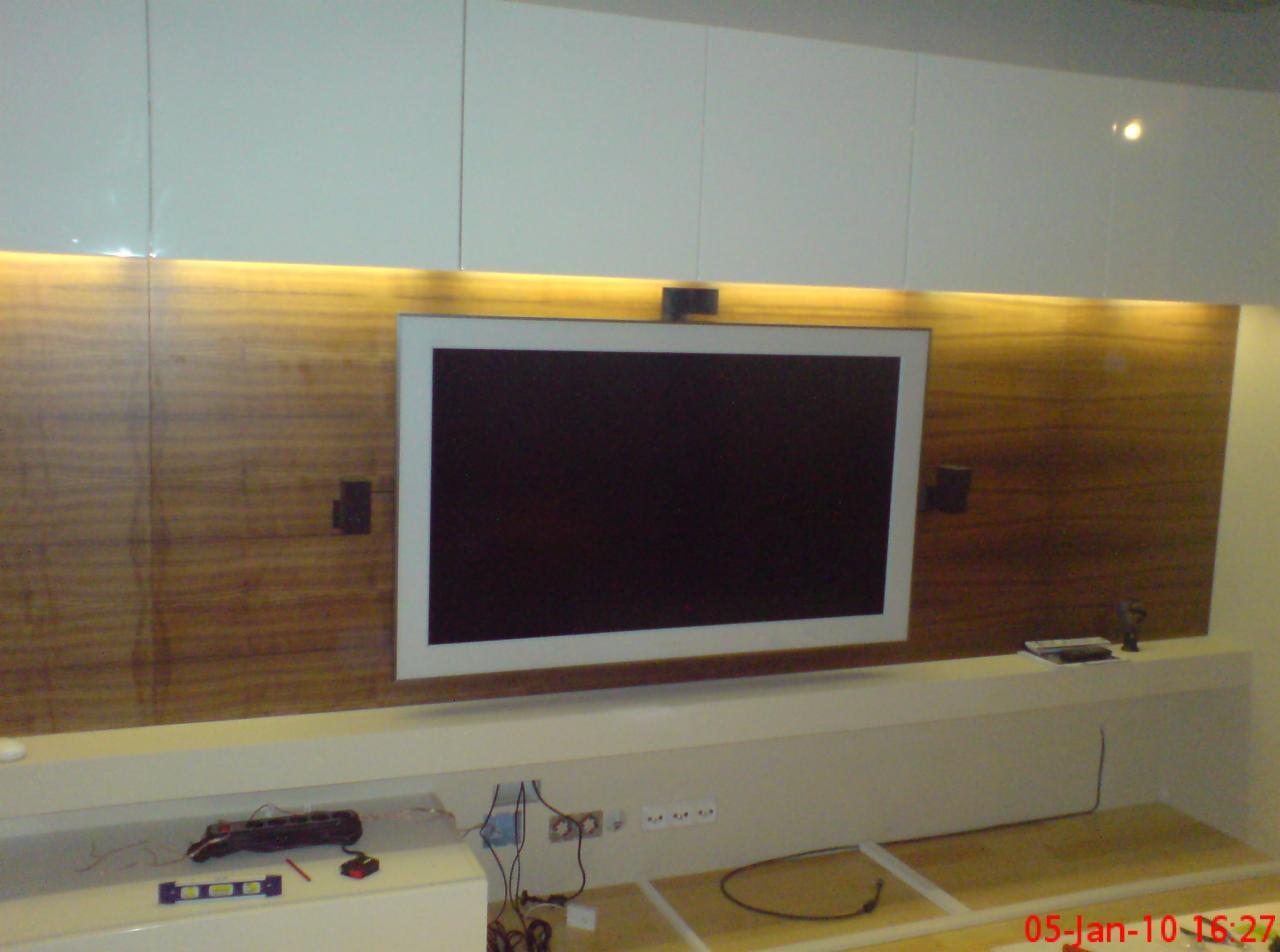 Telewizor z białym obramowaniem zamontowany na drewnianej ścianie z podświetleniem LED, widoczne gniazdka elektryczne i kable w trakcie podłączania.