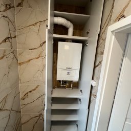 P.P.U.H Dariusz Rogowiec - Biała szafa wnękowa z otwartymi drzwiami, ukrywająca piec gazowy Vaillant, widoczne rury, półki i marmurowe płytki na ścianie.