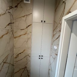 P.P.U.H Dariusz Rogowiec - Wysoka, zabudowana szafa wnękowa z czterema drzwiami i czarnymi uchwytami, w łazience wykończonej płytkami imitującymi marmur z brązowymi żyłami, widoczna kratka wentylacyjna na górze.