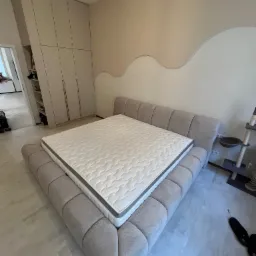 Beżowe łóżko tapicerowane z materacem w sypialni z dużą szafą wnękową i dekoracyjną ścianą w tle.