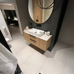 Nowoczesna łazienka z drewnianą szafką pod umywalką, białą umywalką nablatową, okrągłym lustrem z czarną ramą i czarną armaturą, widok z perspektywy żabiej