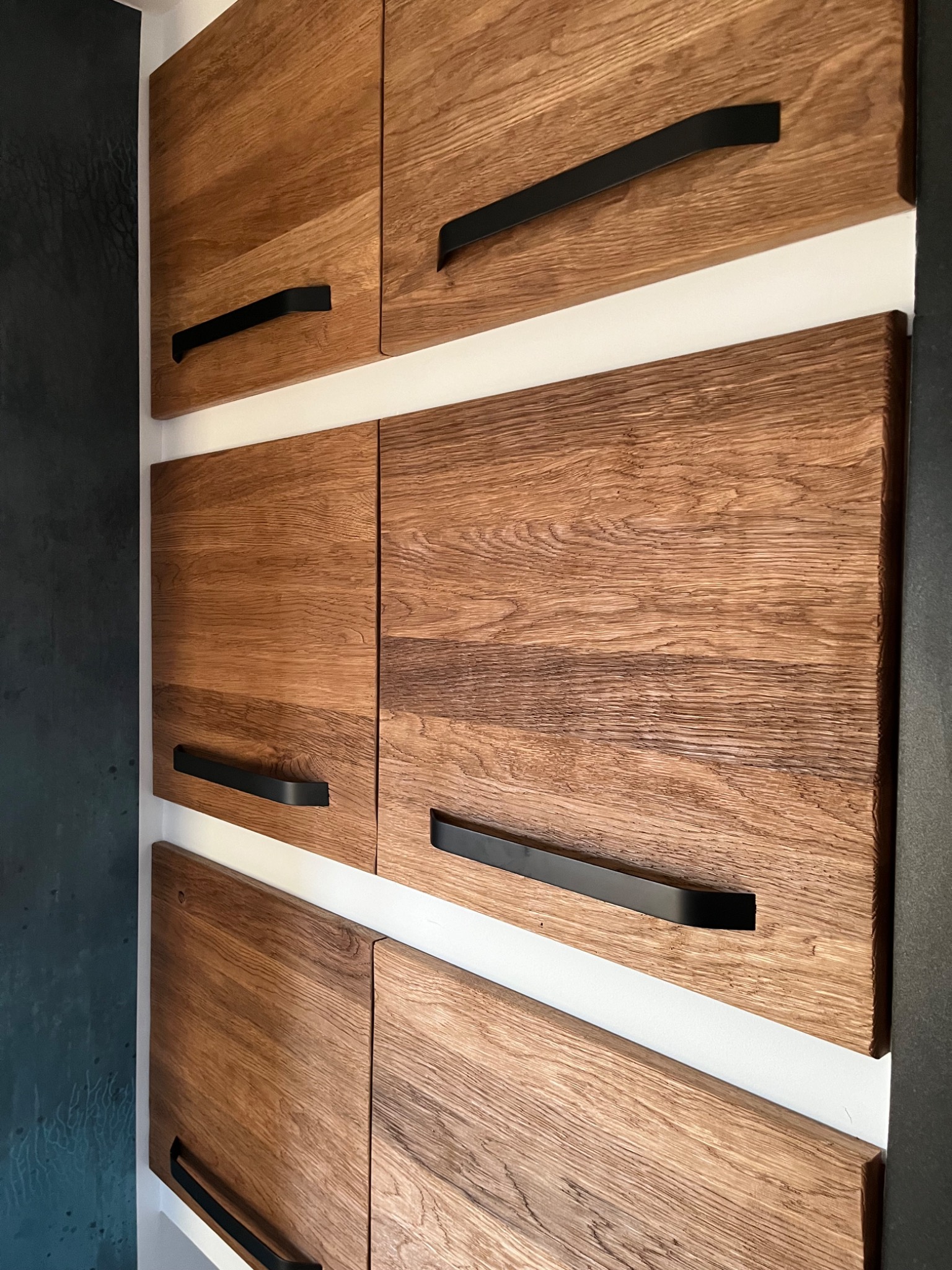 Szczegółowe ujęcie drewnianych frontów szafek z czarnymi, minimalistycznymi uchwytami, oddzielonych białymi elementami, z fragmentem ciemnej ściany w tle.