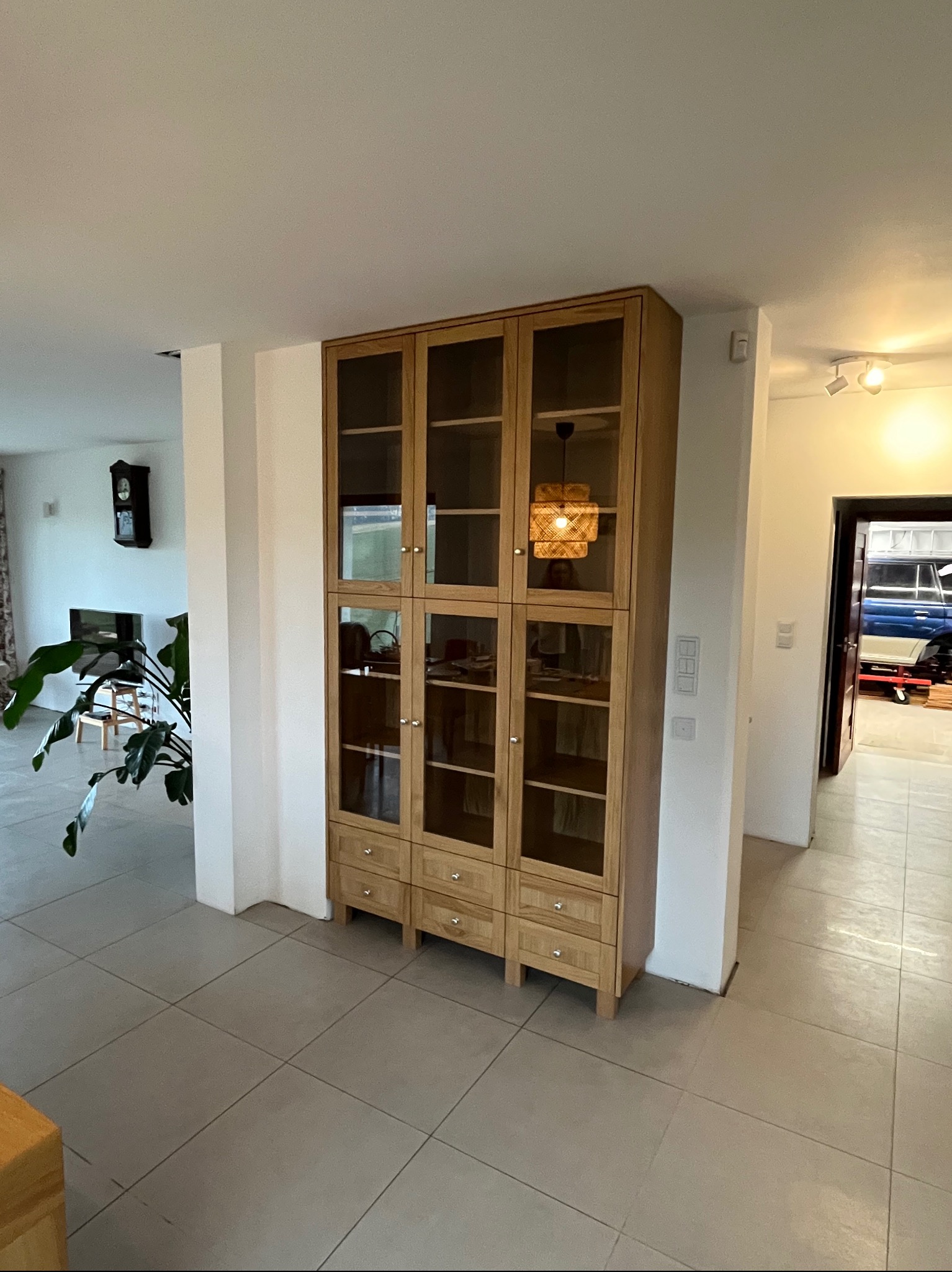 Drewniana witryna z przeszklonymi drzwiczkami i szufladami w salonie, widoczna lampa z abażurem z naturalnych materiałów i fragment wnętrza z rośliną doniczkową.