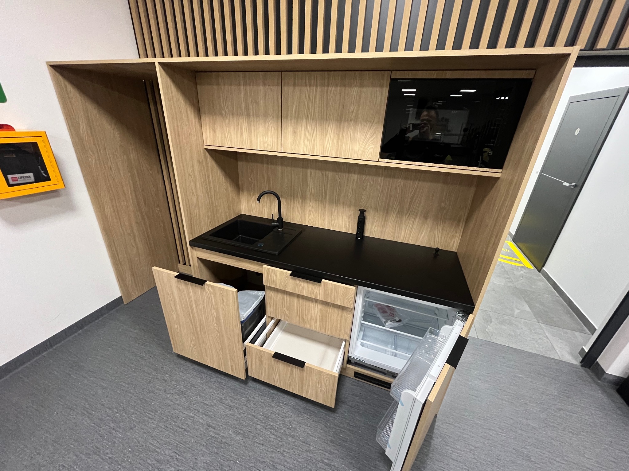 Kompaktowa, drewniana kuchnia z czarnym blatem, zintegrowanym zlewem, mikrofalówką i małą lodówką, umieszczona w biurze lub innym pomieszczeniu socjalnym.