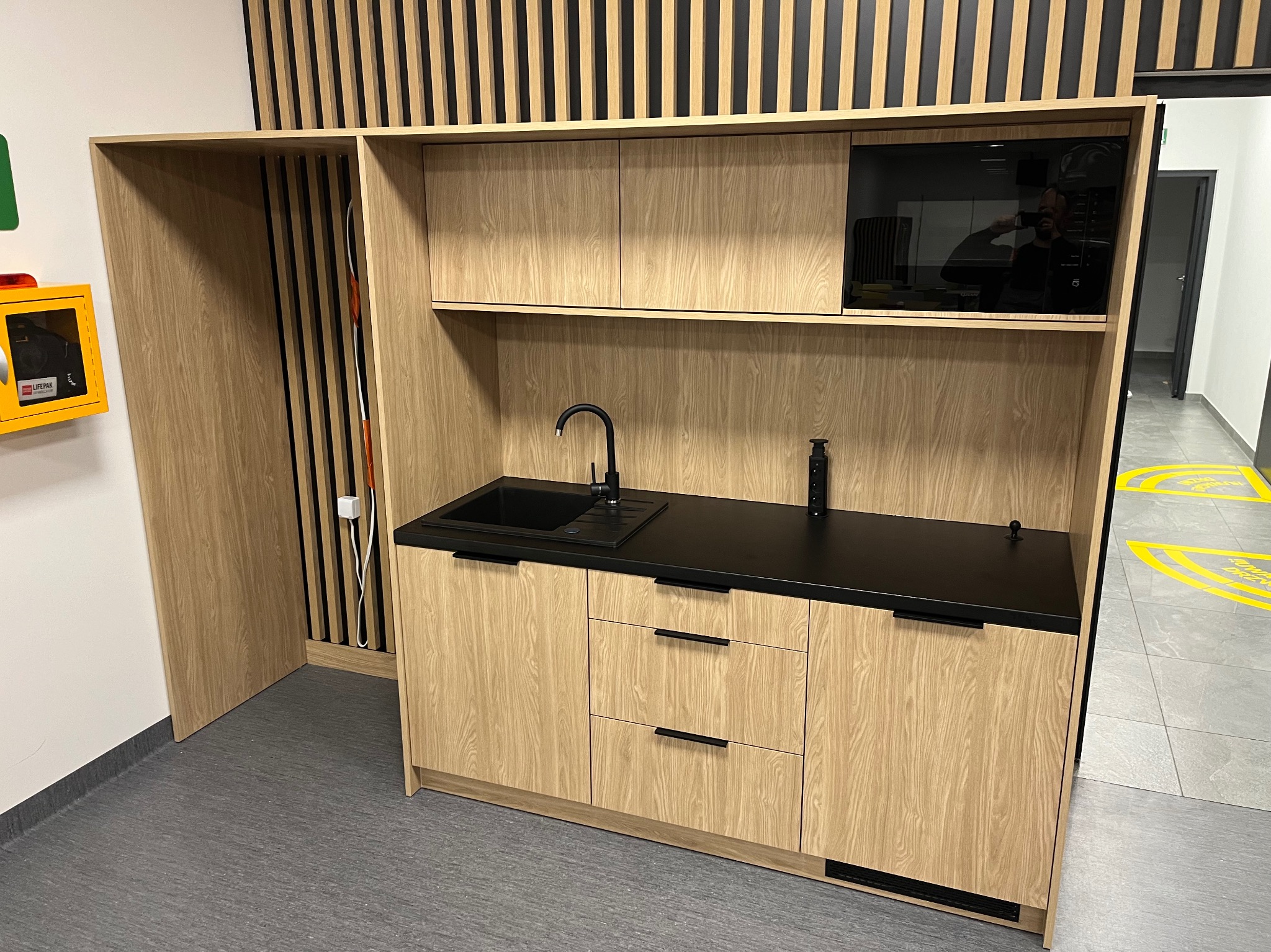 Kompaktowa zabudowa kuchenna z drewnopodobnymi frontami, czarnym blatem i czarną armaturą, uzupełniona o mikrofalówkę i dekoracyjne pionowe lamele na ścianie.