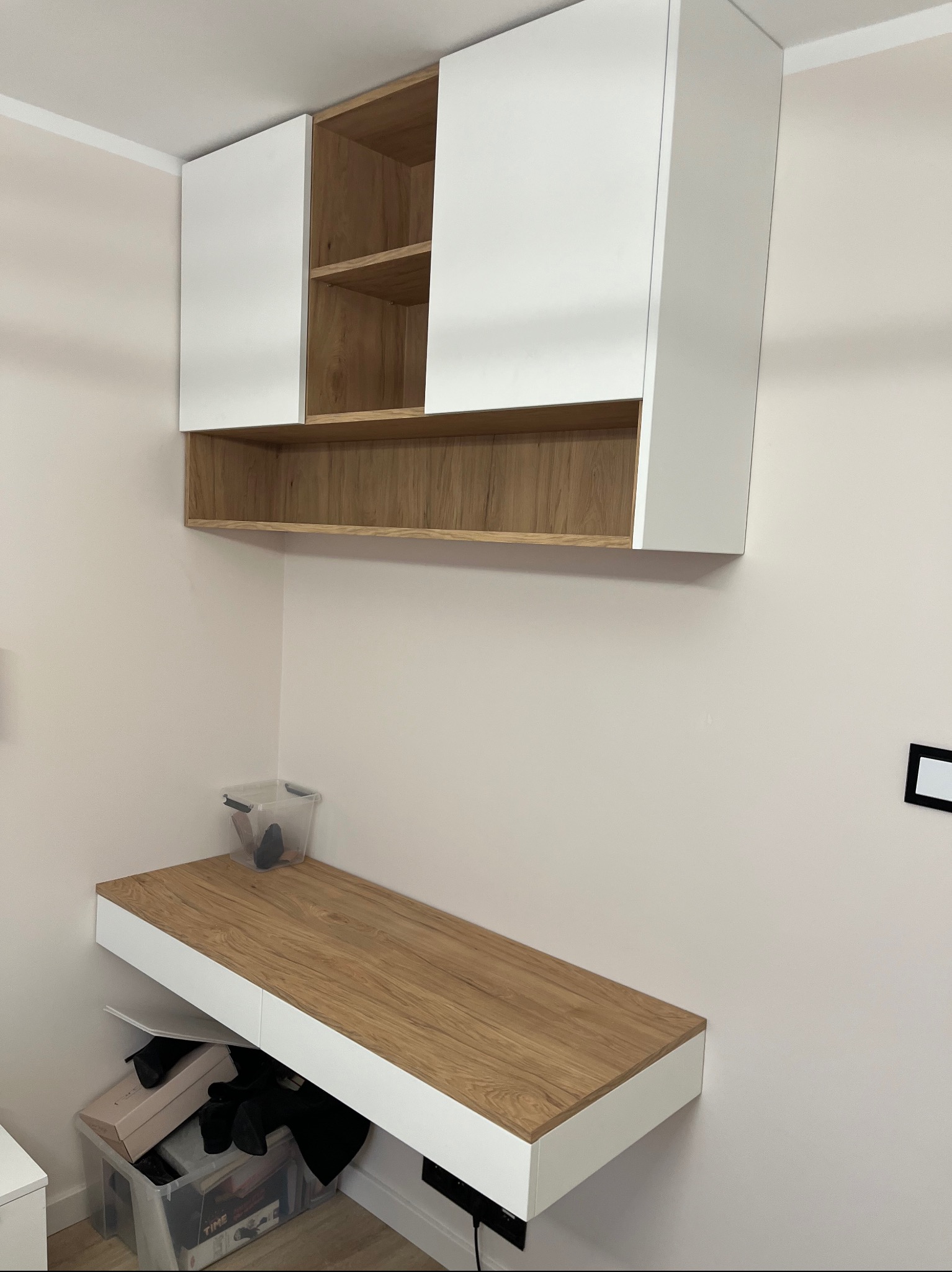 Wiszące szafki kuchenne z białymi frontami i drewnianymi półkami nad blatem biurka z podobnym drewnianym wykończeniem, minimalistyczny design.