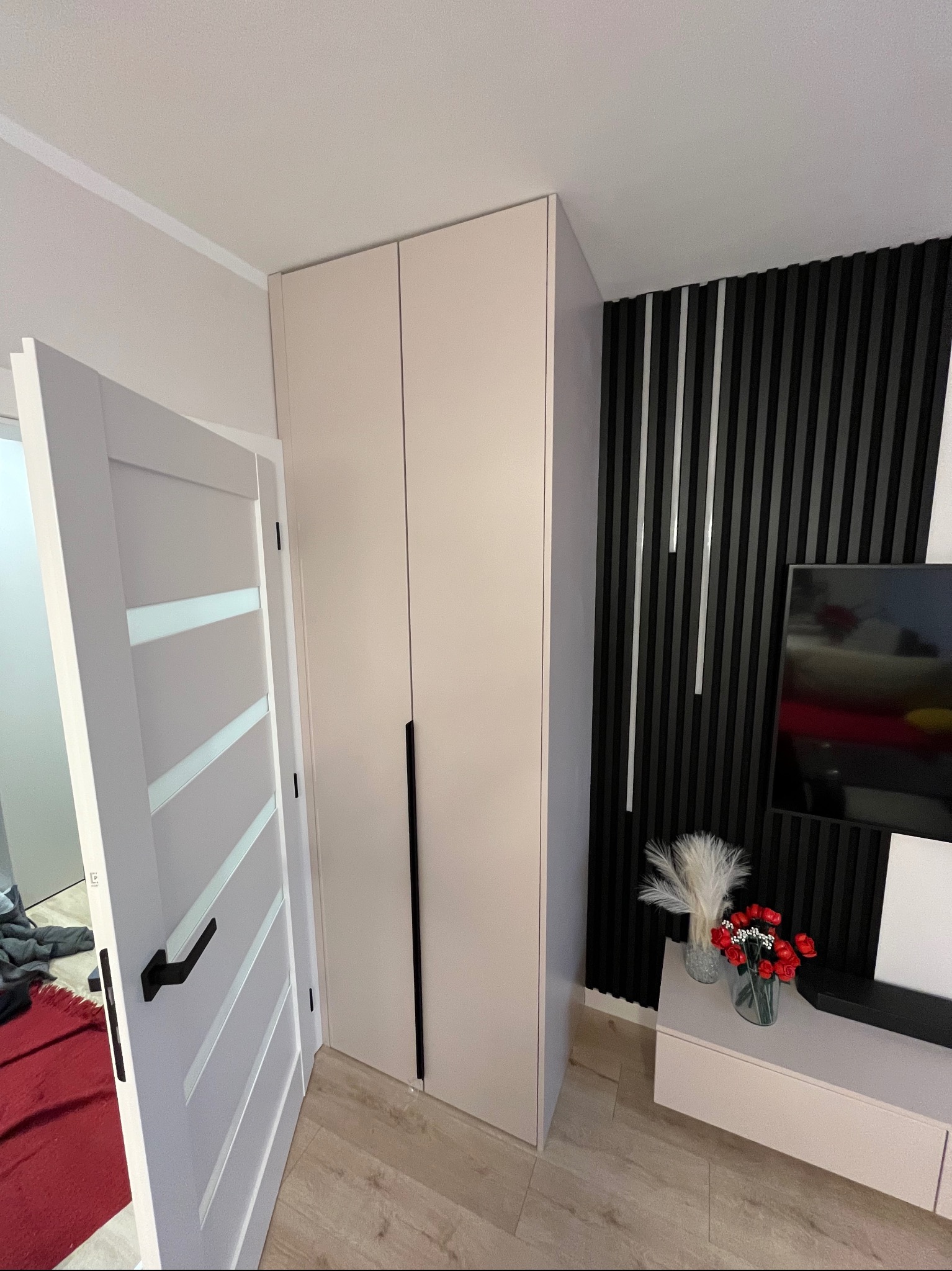 Wysoka, beżowa szafa wnękowa z czarnymi, minimalistycznymi uchwytami, dopasowana do ściany z dekoracyjnymi, czarnymi lamelami i telewizorem, widoczna otwarta biała drzwi z czarną klamką...